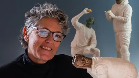 Angus Stewart A woman with short, curly grey hair smiles at the camera. She is wearing black rimmed glasses and a black rollneck top. Some sculptures of skeletons wearing white clothing can be sent to the right of her as you look at the image.