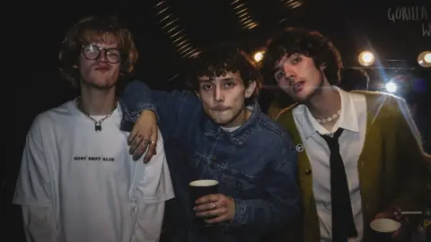 Adora Mekuleyi Luke Hare, Harry Gray, Sol Abrahams from Bilk. Luke, Harry and Sol are all white men in this 20s with grown-out curly hair. Luke has fair hair and a short beard and wears round black-rimmed glasses and a white T-shirt. Harry, a bit shorter than Luke, has brown hair and wears a denim shirt buttoned to the top. Sol tilts his head towards Harry and wears a green cardigan over a white shirt and black tie. The band is pictured inside at a night-time event with drinks in their hands