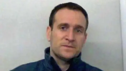 Thames Valley Police Police mugshot of James Sheen. He has short dark hair and is wearing a blue top with the collar up.