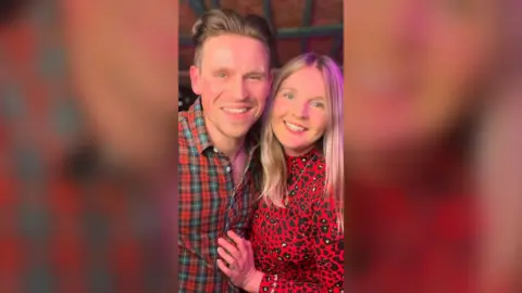 Man and woman both with clone hair smiling. The man is wearing a red checked shirt the woman is wearing a red top with black spots.