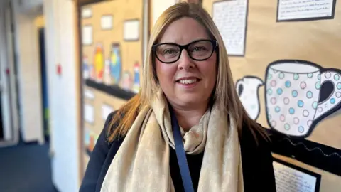 Hope Rhodes / BBC Head teacher Sarah Clowes is looking straight into the camera in the middle of the picture, in front of a wall display in a school corridor. She is smiling, has straight blonde hair and wears glasses, as well as a golden scarf.