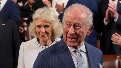 Getty Images King Charles smiles, wearing a dark blue pin stripe suit jacket, white shirt and bluje patterned tie, behind him Queen Camilla wears a white top with black stitching around the collar.
