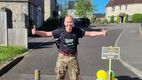 Martyn Harris is standing with thumbs up and smiling at the camera, he is wearing a black t-shirt advertising his Stalbridge 24 hours walking challenge and army trousers.