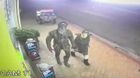 New Zealand Police CCTV still shows a masked and camouflaged man carrying Mr Phillips and one of his children walking down a street in the town of Piopio in November 2023