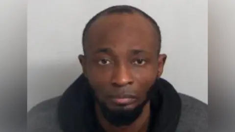 Custody image of Muritala Olaiya-Imam - he is wearing a dark grey top with a thick black collar and looks directly into the screen