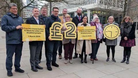 A view of leaders and campaigners in Greater Manchester. They are holding signs about free bus travel for older and disabled people from 1 March 2026, and the £2 fare freeze.