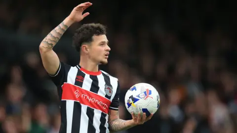 George McEachran, in black and white Grimsby kit, holds up his right arm with the ball held in his left hand.
