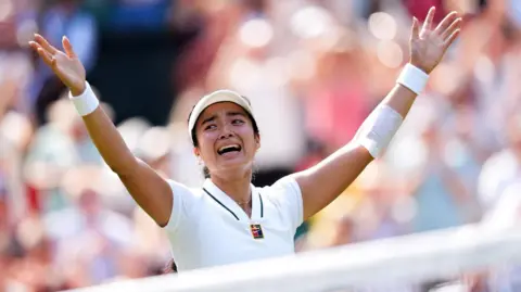 Filipina tennis player Alexandra Eala raises his arms after a Wimbledon match, net in front of her and a blurred image of the crowd in the background