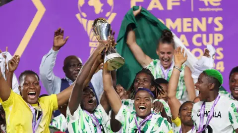 Several members of Nigeria's victorious team lift the trophy after winning the 2024 Women's Africa Cup of Nations. The outfield players are in jerseys with green and white vertical stripes, while goalkeeper Chiamka Nnadozie is seen on the bottom left in a yellow top. A purple board with gold lettering is seen in the background