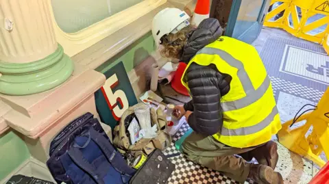 Newcastle City Council View of an artist kneeling down and has just finished painting a small square sign - the number 5 in yellow against a green background. It is leaning against a wall, with brushes and paints alongside it. He is wearing a hi-viz jacket and helmet.