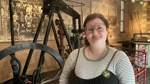 A woman, wearing glasses and a striped top stands in front of an old farming machine.