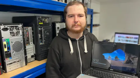 Billy holds a laptop PC showing the Windows home screen as he stands in a workshop in front of shelves full of desktop computers. He has ginger hair and a short beard and is wearing a black hoodie.