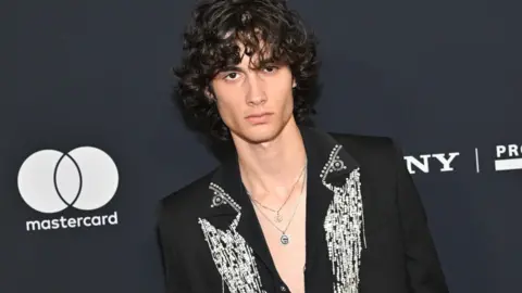 Sombr wearing a black jacket with ornate silver detailing stands before a dark event backdrop featuring Mastercard and Sony branding.
