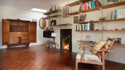 Historic England A view inside Holst's living area inside her home. A fireplace can be seen as well as a nearby wooden chair. A large oak cupboard also sits upon one of the room's walls with its doors open. Bookshelves line the walls.