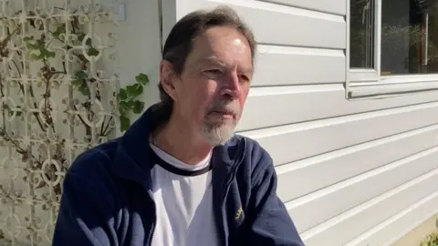 A white man in a white shirt and a navy fleece with a beard and long hair, sits in front of a white house, with a green garden and blue skies. 