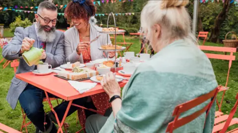 Thrive An elderly woman wearing a pale green shawl and with her back to the camera is sitting at a red picnic table in a large garden. She is joined by a man, looking smart in a grey jacket, tie and jeans, and a younger lady with curly dark hair and a red polka dot dress. There is a cake stand with different cakes on it and the man is pouring himself a cup of tea from a green tea pot. He and the young lady are both smiling. The man has a very large grey beard.