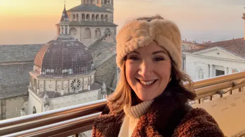 Monica Stott Monica Stott em frente a uma catedral em Bergamo, Itália. Ela está sorrindo para a câmera e vestindo um chapéu creme e casaco marrom. 