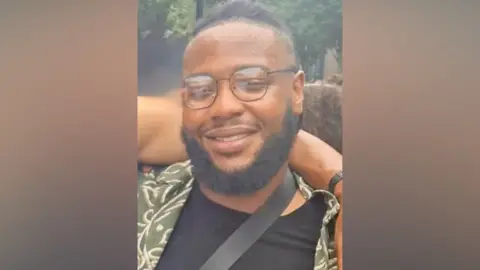 Met Police Close-up portrait of a smiling person with glasses and a beard, wearing a patterned shirt and black top, outdoors with trees in the background.