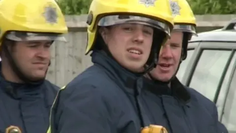 Martyn Sadler stands in firefighter kit wearing a yellow helmet with a visor and a dark blue coat