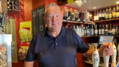Sean Murphy in a dark blue polo shirt stands behind a bar counter, one hand resting on the counter and the other holding a beer tap handle. Behind them are shelves stocked with bottles of alcohol, snacks, and bar items. A chalkboard menu is visible on the wall to the left.

