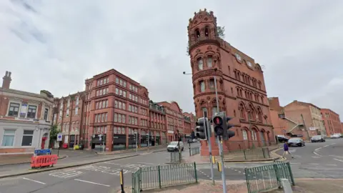An image taken from street level looking up a road with traffic lights in the foreground and old red brick gothic buildings either side of the street. 