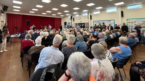 A crowded hall is filled with people sat in rows looking at a stage with a red curtain across it.