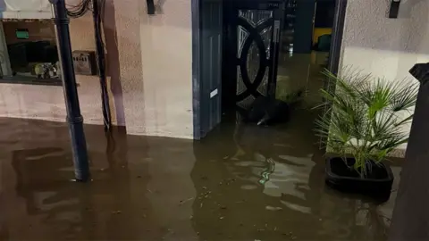 Salique Meah Floodwater covers the ground outside and extends through an open doorway into a building. Muddy water surrounds a potted palm plant beside the entrance, while debris and reflections are visible on the flooded surface.