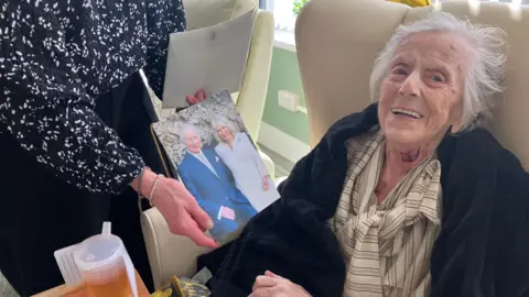 A woman is seated in a high‑backed, light‑coloured armchair upholstered in what appears to be leather or faux leather. The person is positioned slightly to the right side of the frame and faces the camera directly. She has short, light grey hair and is wearing a light‑coloured, buttoned shirt with narrow vertical stripes. A dark, soft-looking garment or blanket is draped around the shoulders and upper arms. She is smiling and receiving a card with a picture of King Charles III and Queen Camilla on.