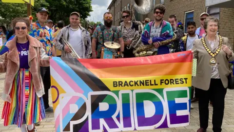 The front of the parade procession. A large Pride banner is carried by two women. One is wearing a long rainbow skirt and a purple t-shirt bearing the Pride logo. The other is wearing mayoral chains. Behind them is a brass band.