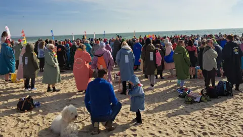 Hundreds of people are standing on a beach with their backs to the camera. They are wearing multi-coloured swimming robes and are facing the sea.