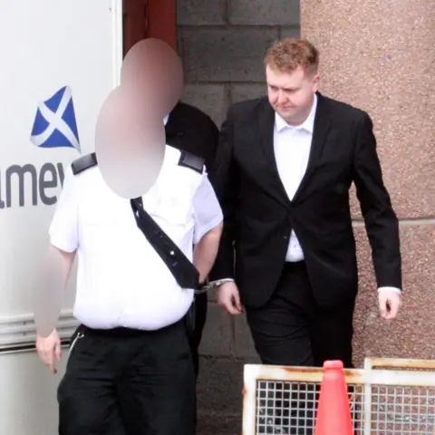 Central Scotland News Agency A man with red hair and a suit is led in handcuffs to a prison van by two guards