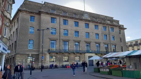 Exterior of the Guildhall in Cambridge. A large square brick building with large windows and a clock at the top. There are market stalls selling fruit and vegetables opposite and people mill about.