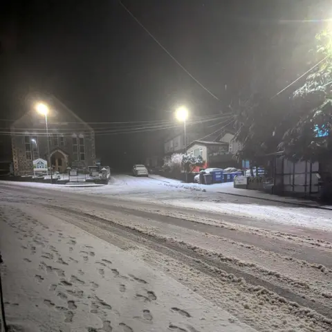Weather Watchers / Paul A street at night, with a series of footprints in the snow. A chapel and nearby houses are lit by street lights. 