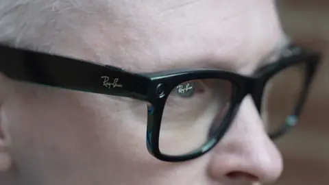 A close up of Clarke's face showing his eyes and nose, with the black glasses. The Ray Ban brand is visible etched on the frames and glass.