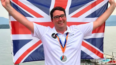 Karl Haines is holding the Union flag behind him whilst wearing his GBR Blind Sailing white polo shirt with a Silver Medal around his neck.