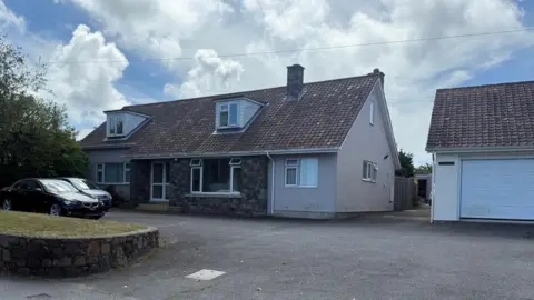 BBC A house, there are two cars in the driveway. There is a green bush to the left. The sky is blue and filled with white clouds. The driveway is grey. 