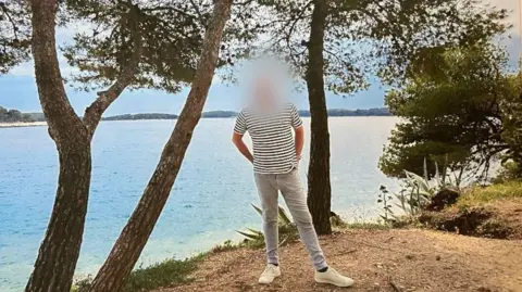 Handout Image shows a antheral   successful  airy   jeans and a striped t shirt. His look   has been blurred out. He is stood successful  beforehand   of a lake, surrounded by trees. The entity  is bluish  and helium  has his hands successful  his backmost  pockets, looking relaxed.