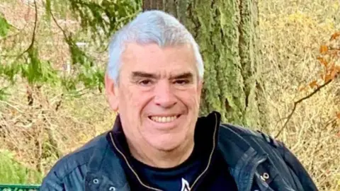 A smiling man in his early 60s with silver hair, wearing a black leather jacket, sitting outside on a bench