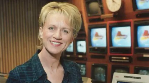 An archive photo of a smiling Sally Taylor posing for the camera in a studio gallery. Behind her is a bank of televisions on the wall.