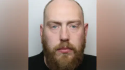 A police custody photo of Joseph Hewitt. He has receding red hair which is cropped short and a red beard. He is wearing a black top.