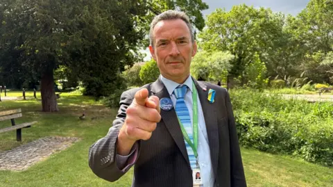 Ed Gemmell in a suit and blue striped tie stands in a park, pointing directly at the camera, wearing a green lanyard and a “Climate Party” badge, with trees, grass and a bench in the background.
