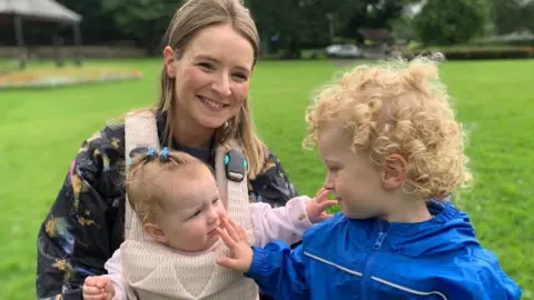 Catherine Burns smiles into the camera. She has shoulder length fair hair and is wearing a black coat with purple and yellow patches. She has a baby harness attached to her chest which is holding baby Maggie. Maggie has three blue hair clips in her blonde hair. She is looking at her two year old brother Leo who has blonde curly hair and is wearing a blue coat. The children and looking at each other and touching each others faces. They are sitting in a grassy area.
