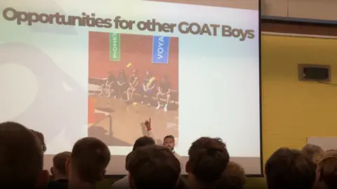 A man standing in front of a large screen saying 'Opportunities for other GOAT Boys'.