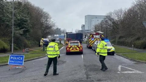Two police officers standing at a junction of a road. There is a blue police sign to the left of the image and there is a fire car, a police car and a fire engine in front of them. 