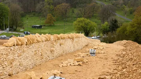 National Highways A golden dry stone wall is pictured on sand with a heap of stone to the right. In the distance are cars, trees and a steep road on a green hill.