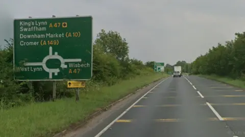 Google A dual carriageway is going towards a roundabout. There is a sign on the left which shows the roundabout, and the road is surrounded by trees and hedges.