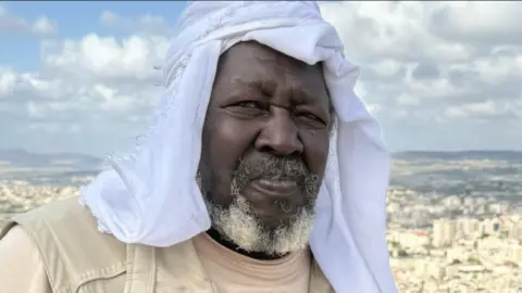 A man wearing headdress looks at the camera with a far off background behind him 