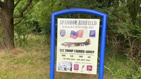 Qays Najm/BBC A sign about Spanhoe Airfield explaining it was a World War Two USAAF station held in a bright blue frame with trees behind