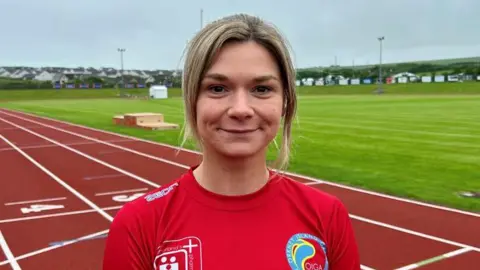 Katie Cubbon Katie Cubbon wears a red T-shirt and stands by a red running track with a green patch of grass in the centre. She smiles and has fair hair which is tied back.
