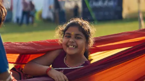 Andrew Prod A young girl is sitting up in an orange hammock. She is wearing a necklace and her dress is embroidered. She is smiling.